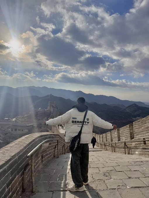 A person in a white 'GEAROTY' puffer jacket standing with arms outstretched on the Great Wall of China, facing a scenic view of mountains and dramatic sunlit clouds.