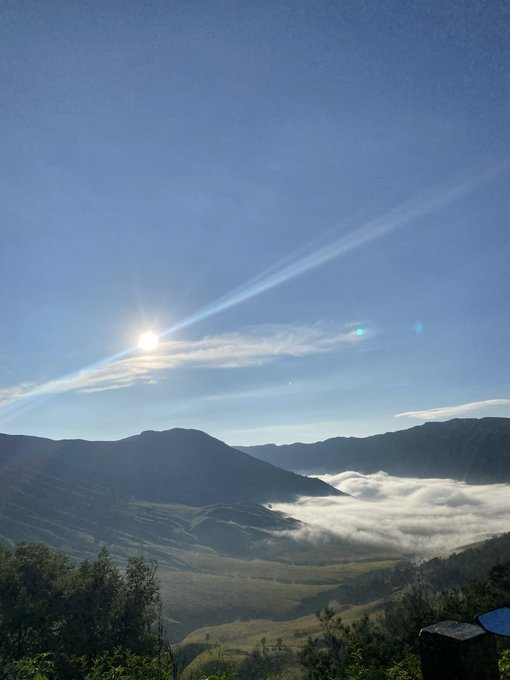 A serene landscape photograph shows a mountain valley filled with a thick sea of low clouds, illuminated by the sun shining brightly through wispy cirrus clouds in a clear blue sky.