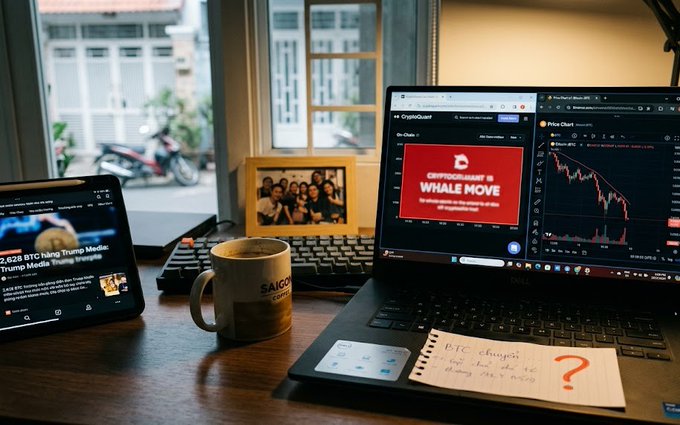 A workspace desk setup featuring a laptop displaying crypto charts and a 'CryptoQuant WHALE MOVE' graphic, alongside a tablet, coffee mug, and a notepad with 'DTC' written on it.