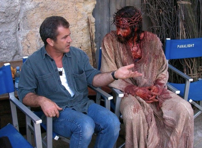 Mel Gibson sits in a director's chair next to an actor in full crucifixion makeup and costume on a film set.