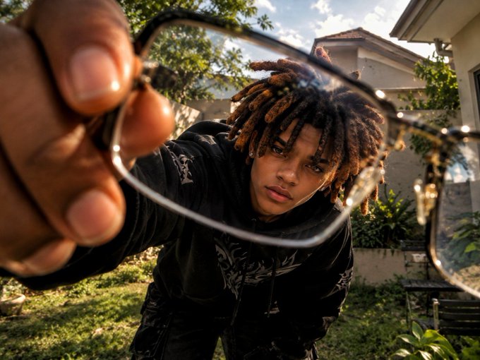 A young man with dreadlocks wearing a black hoodie holds a pair of glasses up to the camera, looking through the lenses from a low angle in a sunny outdoor setting.