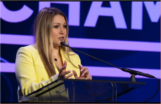 A woman in a yellow blazer speaks at a podium with the partial word "CHAM" visible on a purple backdrop behind her, gesturing with her hands as she addresses an audience. The image, paired with the post about the CLARITY Act's legislative progress, suggests she may be a speaker or advocate discussing the bill's developments while Congress works to reconcile committee texts before the July 13 recess.