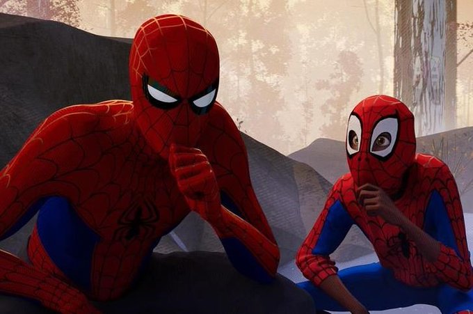 Two versions of Spider-Man sit together in a forest setting, one with the traditional black webbed mask and the other with larger expressive eyes, in a scene from the animated film Spider-Man: Into the Spider-Verse.