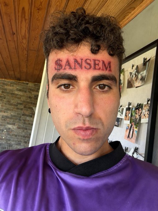 A young man with curly hair and a purple shirt shows off a fresh "$ANSEM" tattoo freshly inked across his forehead, marking the bold, unpaid tribute referenced in the post about Spanish influencer StarPlatinum.