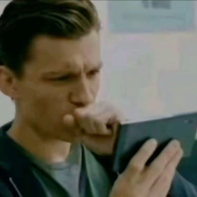 A close-up of actor Tom Holland appearing to cough into his fist while holding what appears to be a tablet or phone.
