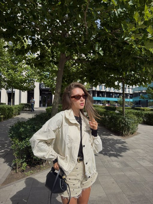 A woman wearing sunglasses, a white jacket, and a skirt poses on a sidewalk lined with green trees and bushes in an urban setting.