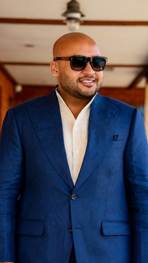 A bald man wearing tortoiseshell sunglasses and a navy blue blazer over a beige mandarin-collar shirt smiles slightly, exuding a confident, polished demeanor that fits the "massive" Bitcoin theme.