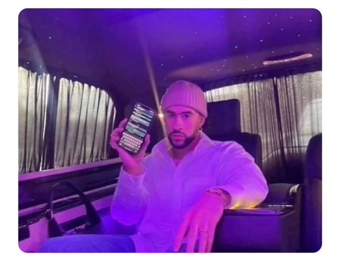A man in a pink beanie and white shirt sits in a vehicle with purple lighting and a starry ceiling, holding up a smartphone displaying a chat interface.