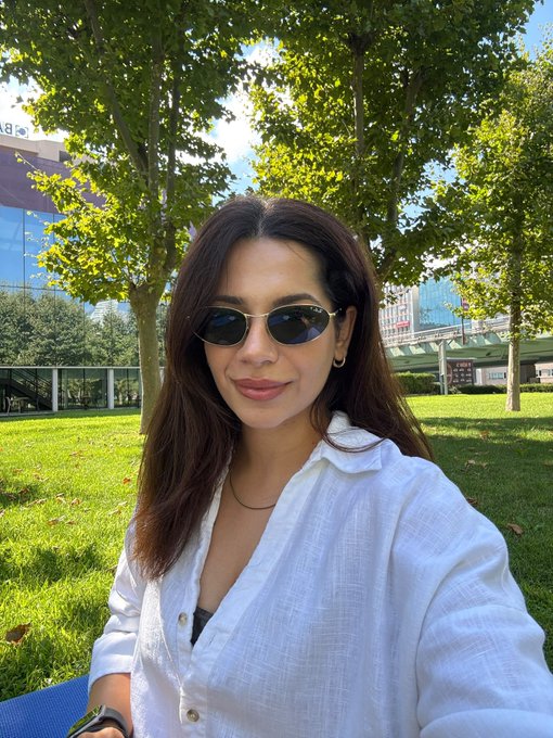 A selfie of a woman with long dark hair wearing sunglasses and a white shirt, taken outdoors on a sunny day with trees and modern buildings in the background.