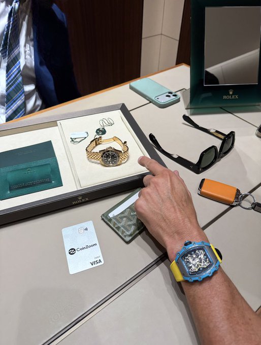 A man wearing a blue and yellow luxury watch points at a Rolex display showcasing watches, sunglasses, and accessories, while a "CoinZoom VISA" card and a green cardholder rest on the counter, promoting the CoinZoom app referenced in the post.