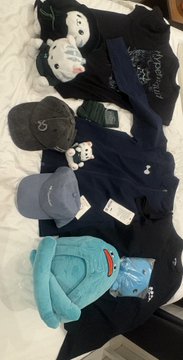 A collection of plush toys and merchandise, including shark-themed items, a Poké Ball, and a blue bunny hat, arranged on a white bedspread.
