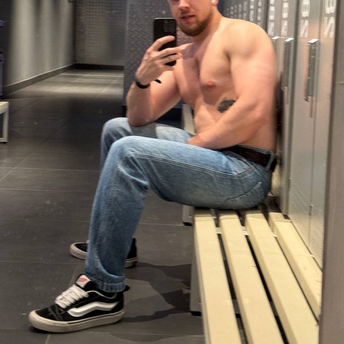 A shirtless, muscular man with a beard takes a mirror selfie while sitting on a wooden bench in a locker room, wearing blue jeans and black Vans sneakers.