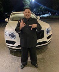 A small-statured man in a black suit posing with hand gestures in front of a white Bentley convertible parked at night.