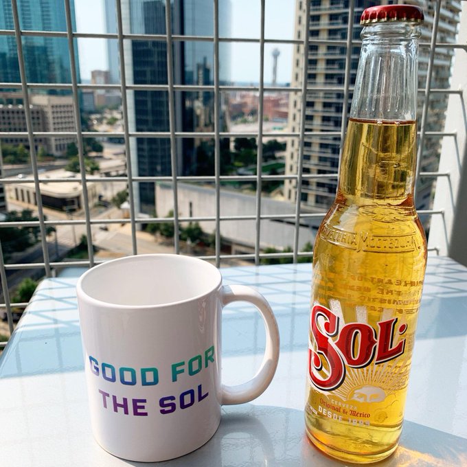 A glass bottle of Sol beer from Mexico sits next to a white coffee mug that reads "GOOD FOR THE SOL," both placed on a glass table overlooking a city skyline through a metal railing.