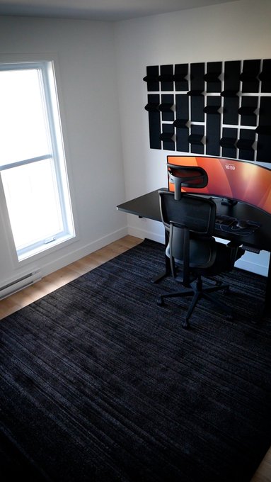 A minimalist home office setup featuring a curved monitor on a black desk, an ergonomic chair, a dark rug, and a wall-mounted acoustic panel grid.