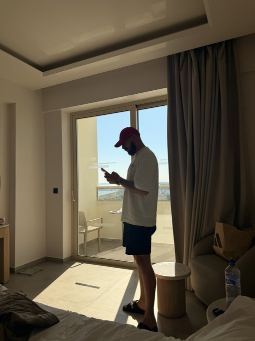 A bearded man in a white t-shirt, dark shorts, and a red cap stands on a sunlit balcony of what appears to be a hotel room, checking his phone. The image likely showcases the luxury accommodation that the "$2,500 giveaway" prize might include, helping to entice participants to engage with the post by liking and retweeting.