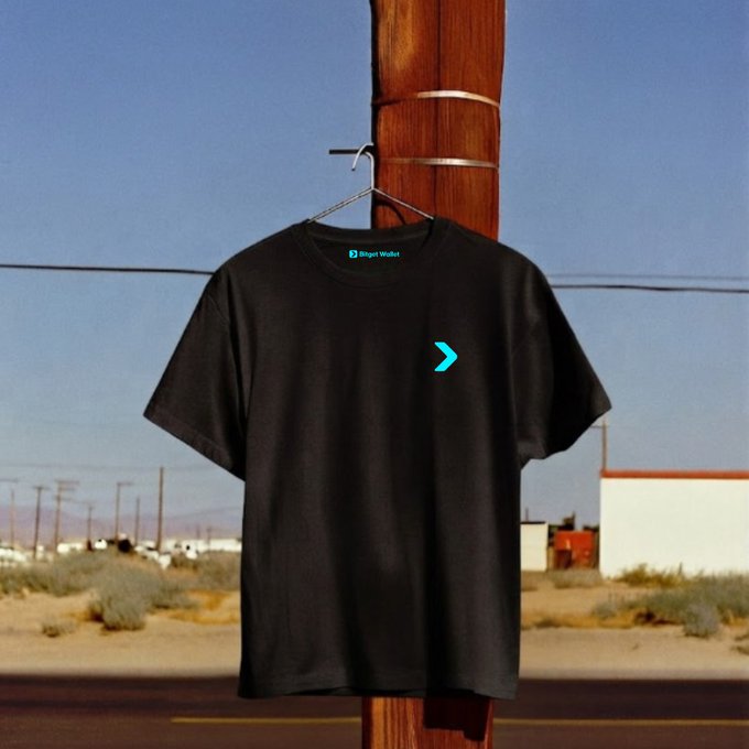 A black t-shirt featuring a teal "B" logo and "Bitget Wallet" text on the chest hangs from a wooden utility pole against a desert backdrop. The casual, laid-back setting complements the motivational Monday message about taking action today without waiting for the perfect moment.