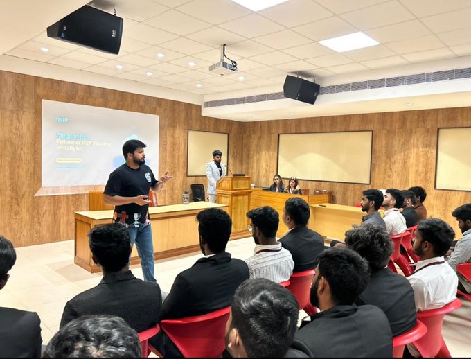 A man in a black t-shirt presenting at a workshop, with a projection screen behind him reading 'Workshop: Technical CSV Trading with Python' by 'mr.tr', while an audience sits in a wood-paneled lecture hall.