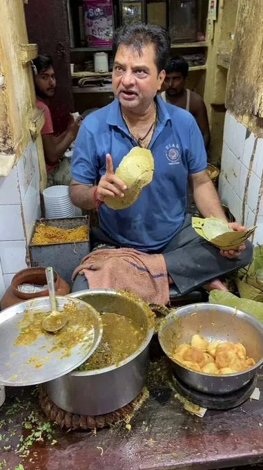 A man in a blue polo shirt sits behind pots of curry and potatoes, peeling what appears to be a large fruit or vegetable with a leaf in hand. The image humorously parallels the "ArcDev cooking new launchpad" post by depicting a literal "cooking" scene, suggesting a playful metaphor for developers preparing a new product launch.