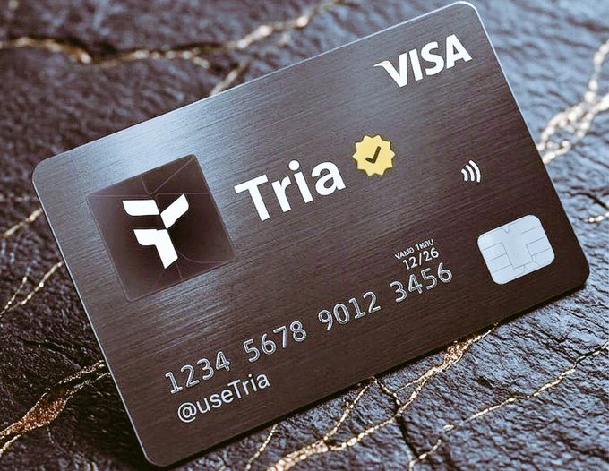 The image shows a black Visa credit card with white text that reads "TRIA" and "useTria In Web3", indicating that the card belongs to someone named Tria who uses the service provided by @useTria. The credit card is placed on a rock, suggesting it might be a souvenir or keepsake from a trip.