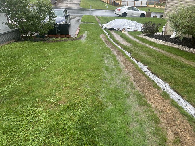 A residential front yard shows a freshly dug drainage trench lined with white landscape fabric running alongside the lawn, with parked cars and a wet driveway visible in the background.