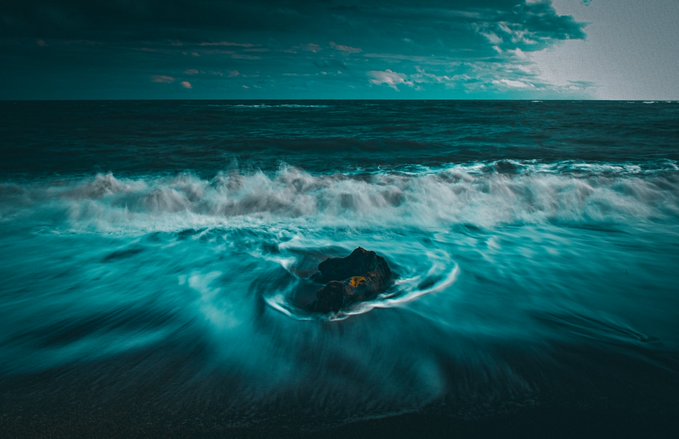 A dramatic seascape with turquoise waves swirling around a dark rock, under a moody teal sky with scattered clouds.