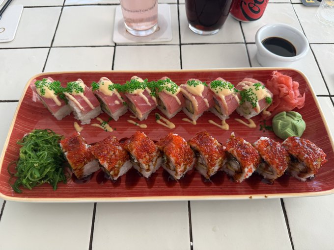 A rectangular red plate of sushi featuring two rows of rolls topped with tuna and fish roe, garnished with green seaweed salad, pickled ginger, and wasabi, with drinks in the background.