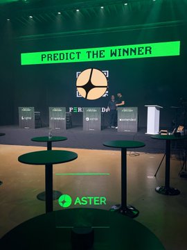 An event stage with green lighting displays 'PREDICT THE WINNER' on a screen, with a person at a podium and empty cocktail tables in the foreground.
