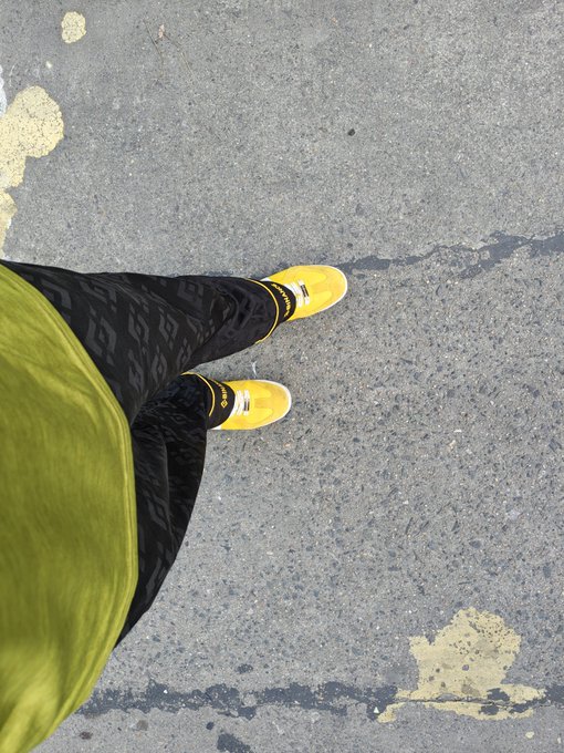 A downward-facing photograph showing a person's legs wearing black patterned pants and bright yellow sneakers with white text, standing on gray asphalt.