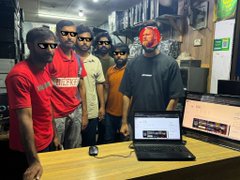 A group of men stand behind a counter in what appears to be a retail store; one wears a red Ferrari polo shirt, another wears a red cap, and a laptop sits open on the counter in front of them.