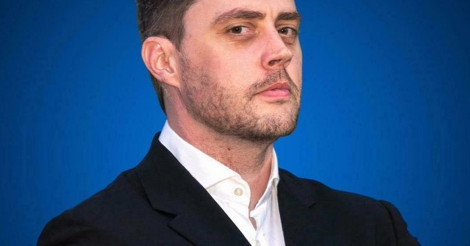 A close-up portrait of a man with short dark hair and stubble, wearing a dark suit jacket over a white shirt against a solid blue background.