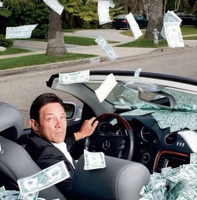 A man in a suit sits in the driver's seat of a convertible car while money bills fly through the air around him on a residential street.