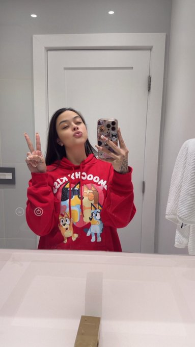 The image features a woman taking a selfie with her phone, wearing a red hoodie that has cartoon characters printed on it. The background includes a bathroom setting with a mirror and a towel hanging nearby. The caption of the photo reads "Happy Sunday to all the employees who have to work tomorrow Day 148 of a selfie a day until MON hits ATH."
