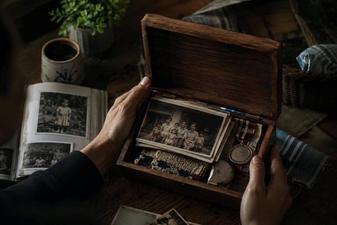 A person holds open a wooden box containing vintage black-and-white family photographs, a pocket watch, and other cherished heirlooms, while an old photo album and potted plant rest nearby—visually evoking the precious family memories and legacy that the post speaks of preserving.