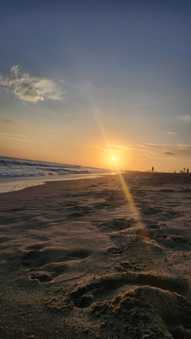 A sunset over a sandy beach with ocean waves on the left, footprints in the foreground sand, and a few small silhouettes of people in the distance.