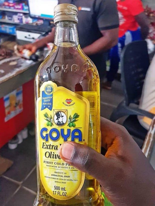 A close-up photo of a hand holding a 500 mL bottle of Goya Extra Virgin Olive Oil, First Cold Pressed, in what appears to be a grocery store or market setting.