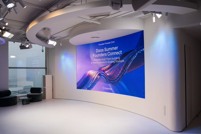 A modern, futuristic room features a large curved LED display screen on the wall showing 'Daos Summer Founders Connect' and 'Founder Chat: From Building in the Trenches to Scaling Startups' hosted by Daos.fun.