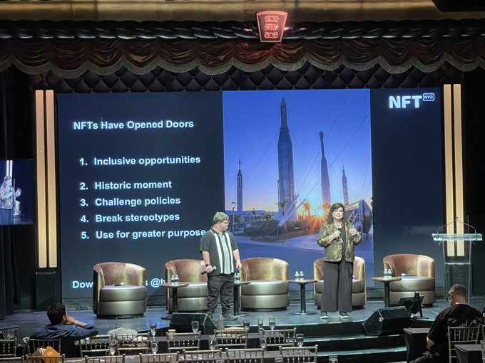 A conference stage with a presentation titled 'NFTs Have Opened Doors' listing points like 'Inclusive opportunities' and 'Historic moment,' with two speakers standing before the screen.
