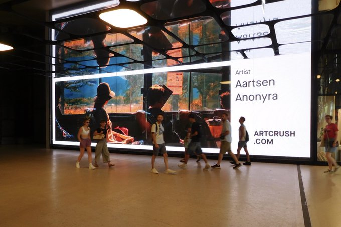 People walking past a large illuminated digital billboard displaying artwork by 'Artist Aartsen Anonyra' with the website 'ARTCRUSH.COM' visible.