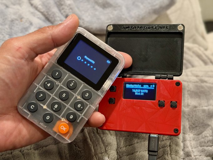 A hand holding a transparent cryptocurrency hardware wallet with a keypad and small screen showing 'SpaceSVM', positioned next to a red 'BOOTYSHACKS' hardware wallet displaying '16.07 SATS'.