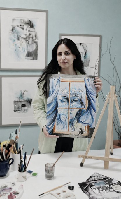 A dark-haired woman in a light blazer holds up an abstract blue and white watercolor painting in an art studio, with framed artworks and an easel visible behind her.
