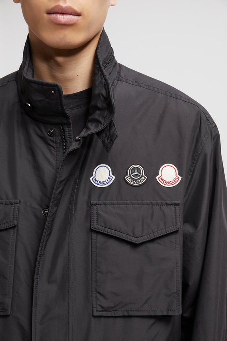 A model wearing a black field jacket featuring three 'Moncler' logo patches (blue, black, and red) on the chest.