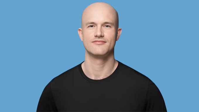 A headshot of a bald man wearing a black t-shirt against a solid blue background, smiling slightly at the camera.