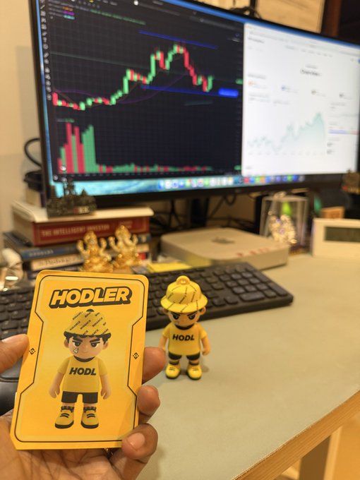 A hand holds a yellow "HODLER" trading card featuring a cartoon character in a hard hat, with a matching figurine on a desk displaying financial charts on a monitor behind it.