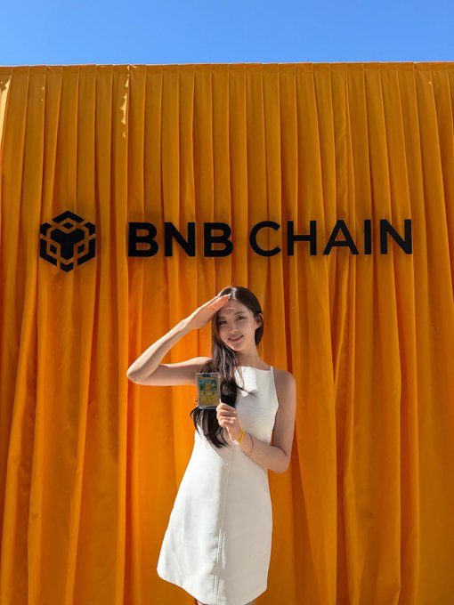 A woman in a white dress salutes while holding a microphone in front of a yellow backdrop with the 'BNB CHAIN' logo.