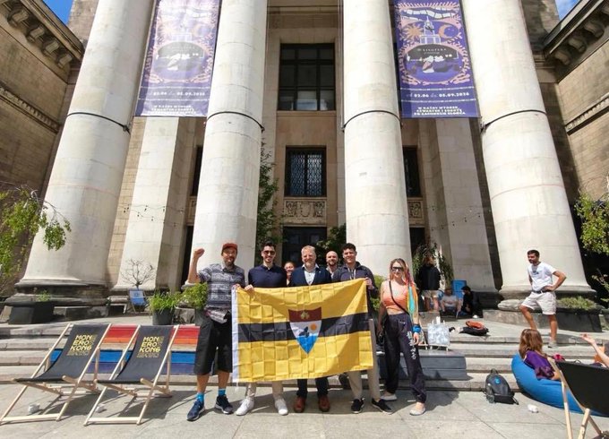 The image features a group of people standing outside an old building with columns, posing for a photo together. The group consists of at least nine individuals, all wearing casual attire such as t-shirts or hoodies. One person among them is holding up a flag that has the Monero logo on it. The backdrop of the scene includes several banners and signs, possibly related to an event or conference.