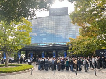 A crowd of people gathered outside a modern glass building surrounded by trees, with a sign partially reading 'XPR' visible on the right.
