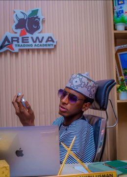 A man in traditional attire and cap holds up a phone with the "AREWA TRADING ACADEMY" logo visible on the wall behind him, celebrating YahayaCrypto's wedding day and the anticipated new all-time high for $SUNUSI.