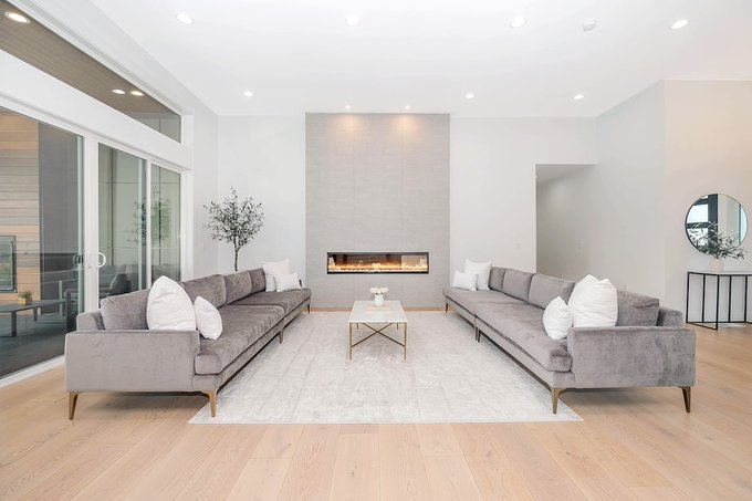 The image shows a spacious living room with two gray couches facing each other, creating an open and inviting atmosphere. The room features a fireplace as well as a coffee table placed between the couches. A large mirror can be seen on one of the walls, adding depth to the space. There is also a potted plant in the corner, contributing to the overall ambiance. The image seems to be related to the post about Monday mornings and how they feel like a fresh checkpoint for building momentum or staying busy throughout the week.