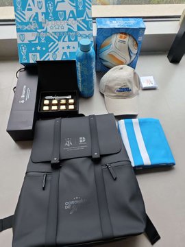 An assortment of promotional merchandise and branded items laid out on a desk, including a gift box, water bottle, cap, backpack, and various printed materials in blue and black branding.
